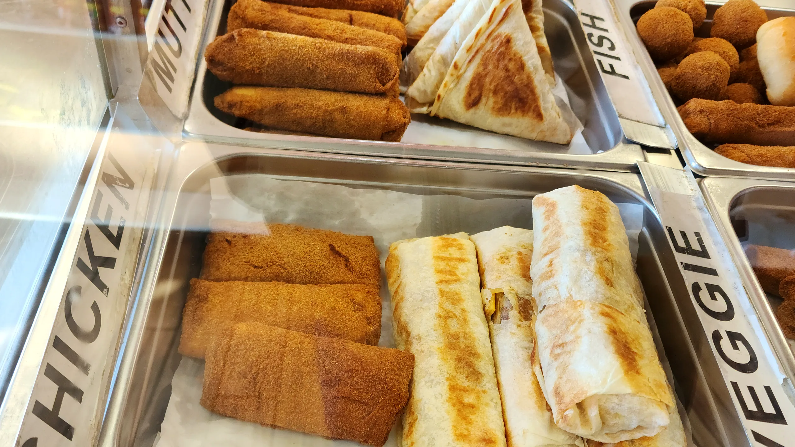 Short eats counter with trays of crumb-fried mutton and chicken rolls, grilled wraps and fish cutlets